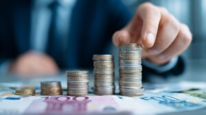 Ruka poslovne osobe slaže uredne stubove kovanica na stolu prepunom evro novčanica, simbolizujući ulaganje malog kapitala u pokretanje biznisa