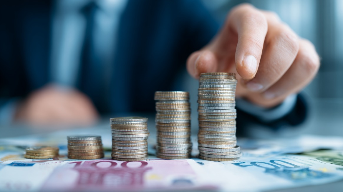 Ruka poslovne osobe slaže uredne stubove kovanica na stolu prepunom evro novčanica, simbolizujući ulaganje malog kapitala u pokretanje biznisa
