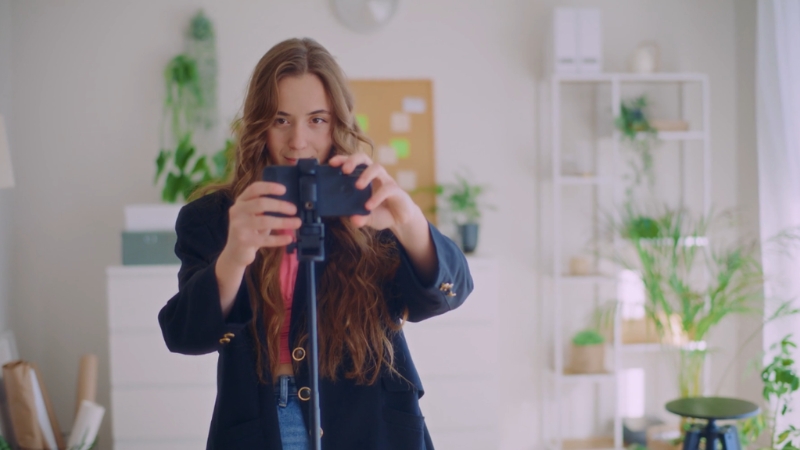 Devojka podešava telefon na stativu u svetloj sobi punoj biljaka i dekoracije